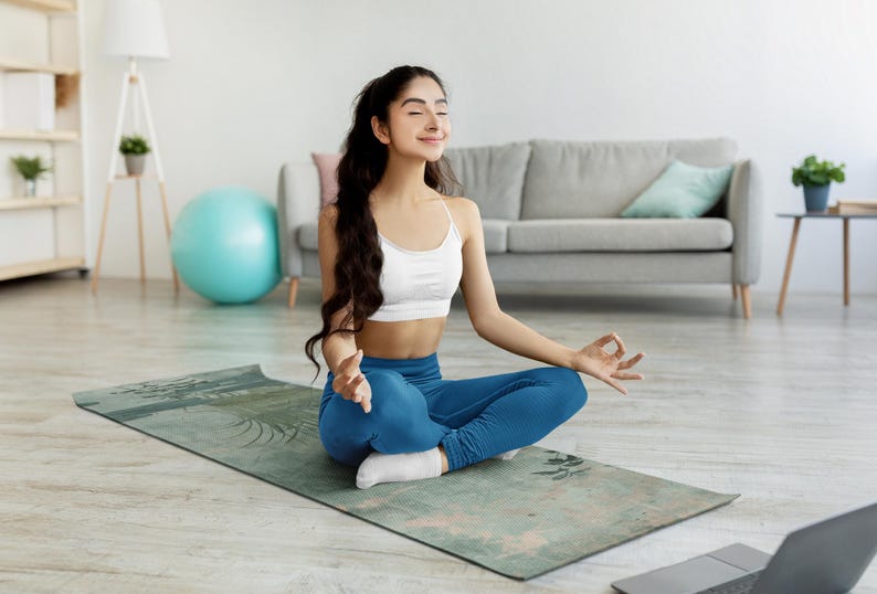 May include: A woman sits in a yoga pose on a blue and green patterned yoga mat. She is wearing a white sports bra and blue yoga pants. She has long dark hair and is looking up with her eyes closed.