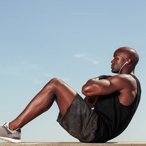 May include: A man wearing a black tank top and camouflage shorts does sit-ups while wearing headphones.