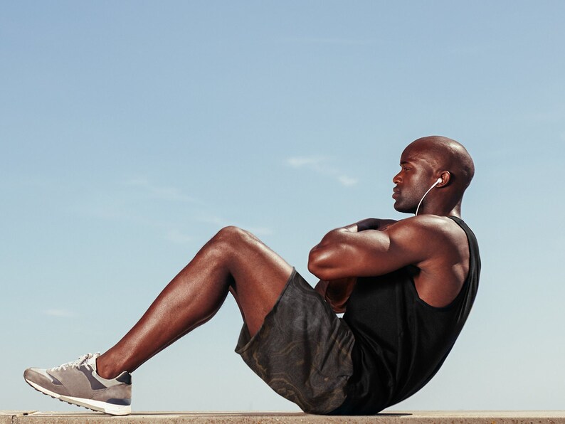 May include: A man wearing a black tank top and camouflage shorts does sit-ups while wearing headphones.