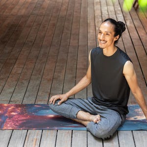 Puede incluir: Una persona sentada en una postura de yoga en una esterilla de yoga colorida con estampado de galaxia. La persona lleva un top negro sin mangas y pantalones grises.