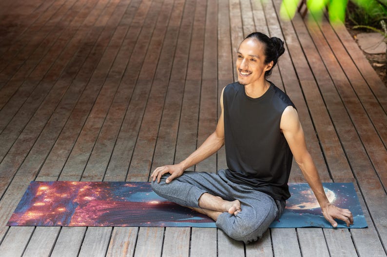 Puede incluir: Una persona sentada en una postura de yoga en una esterilla de yoga colorida con estampado de galaxia. La persona lleva un top negro sin mangas y pantalones grises.