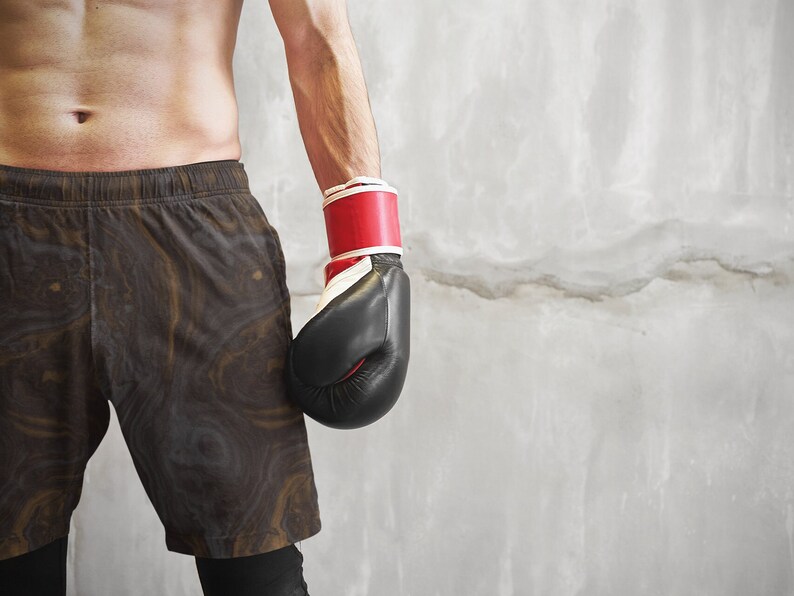 May include: A person wearing black and brown patterned shorts with a red and white boxing glove on their left hand.