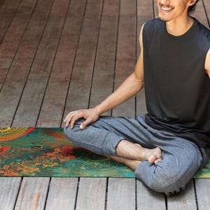 May include: A person sits in a meditative pose on a colorful yoga mat with intricate patterns in shades of green, red, gold, and black. The mat is on a wooden floor.