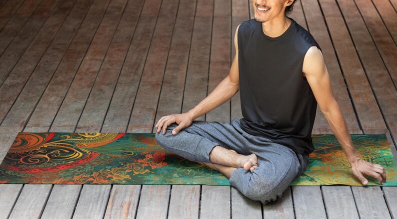 May include: A person sits in a meditative pose on a colorful yoga mat with intricate patterns in shades of green, red, gold, and black. The mat is on a wooden floor.