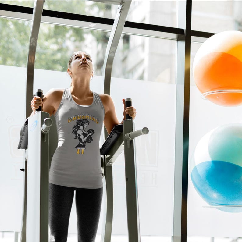 Peut inclure: Un d&eacute;bardeur gris clair avec le texte "Crushing It" et un dessin d'une femme soulevant des poids. Le d&eacute;bardeur est port&eacute; par une personne utilisant un appareil d'exercice dans une salle de sport. Le texte et le dessin sont en noir et jaune.