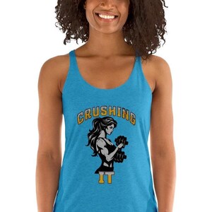 Peut inclure: Un d&eacute;bardeur turquoise avec un graphique d'une femme soulevant des halt&egrave;res. Le texte "CRUSHING IT" est arqu&eacute; au-dessus de la femme en lettres dor&eacute;es et noires. Le d&eacute;bardeur a un design dos nageur.