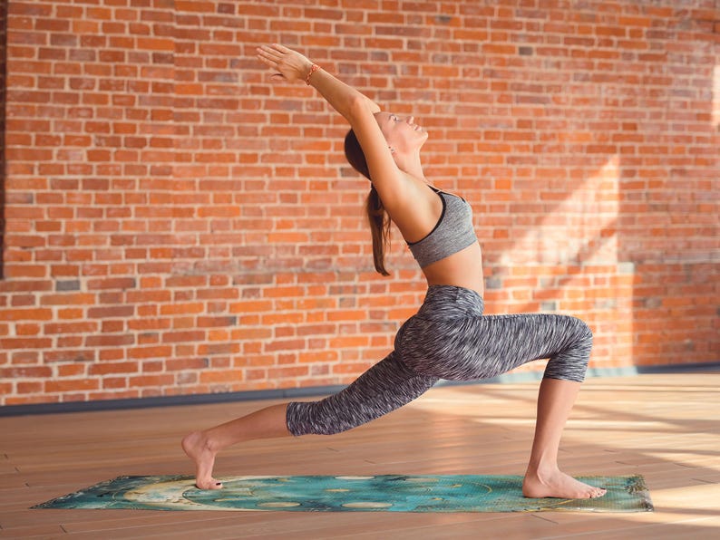 May include: A woman in grey athletic wear performs a yoga pose on a green and blue patterned mat. She is standing with her right leg forward and her left leg straight behind her. Her arms are raised above her head and she is looking up.