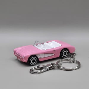 Puede incluir: Llavero de coche descapotable rosa con interior blanco y ruedas negras. El llavero tiene un cierre plateado y un llavero de anilla. El coche tiene una franja blanca en el lateral y la palabra "Chevrolet" está escrita en el lateral.