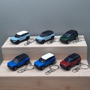 May include: A collection of miniature SUV keychains in various colors, including light blue, dark blue, green, and red. Each keychain features a detailed car model with a black roof and silver keyring attachment. The keychains are displayed on a wooden block.