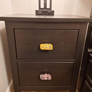 May include: A dark brown wooden bedside table with two drawers. Each drawer has a car-shaped knob. The top knob is yellow with the number 58, and the bottom knob is a red and white car with the number 84.