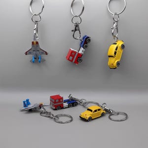 May include: Collection of miniature vehicle keychains. Includes a yellow car, a red truck, a blue and red truck, and a gray airplane. Each keychain has a silver metal ring and clasp for attaching to keys or bags.