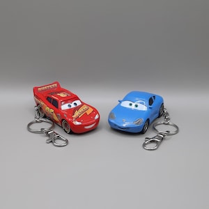 Può includere: Due portachiavi a forma di auto giocattolo. Uno è rosso con il numero 95 e la scritta "RUST-EZE" stampata sopra. L'altro è blu. Entrambi hanno portachiavi e chiusure argentate.