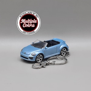 Puede incluir: Un llavero de coche de juguete descapotable azul claro. El llavero presenta un coche en miniatura con interior negro y ruedas plateadas. El coche tiene un llavero y un cierre. El texto "AVAILABLE IN MULTIPLE COLORS OPTIONS" está en un círculo.