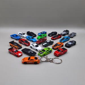 May include: A collection of miniature toy cars in various colors, including red, orange, green, blue, and black. The cars are arranged on a light gray surface. A keychain with an orange car is in the foreground.