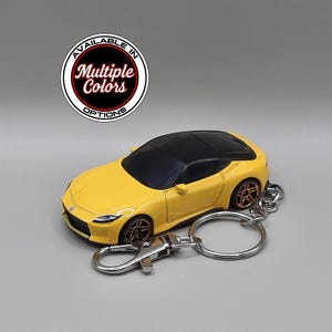 May include: A yellow sports car keyring with a black roof and dark bronze wheels. The keyring features a metal ring and a lobster clasp. Above the car, a circular badge displays the text Available in Multiple Colors Options.