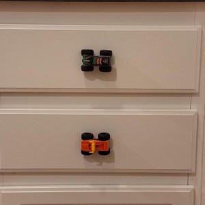 May include: White cabinet drawers with toy monster truck knobs. The top knob is green and black, and the bottom knob is orange and black. The knobs are attached to the white cabinet doors.