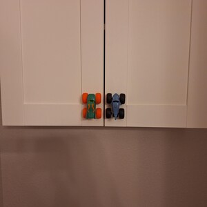 May include: Two cabinet knobs in the shape of toy monster trucks. One is green with orange wheels, and the other is blue with black wheels. They are fixed to white cabinet doors.