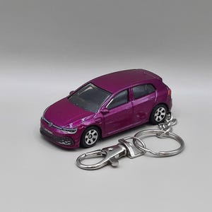 May include: A metallic purple Volkswagen Golf hatchback toy car keyring. The miniature vehicle features black wheels with silver rims, a silver Volkswagen emblem on the front grille, and a silver metal lobster clasp attached by a short chain.