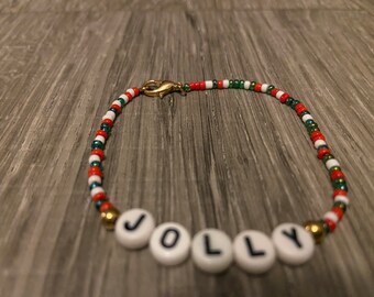 May include: A red, white, and green beaded bracelet with a gold clasp. The bracelet has the word "JOLLY" spelled out in white beads.