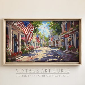 以下が含まれることがあります： アメリカの国旗で飾られたカラフルな建物が並ぶ、趣のある石畳の通りの絵画。通りは空いていて、頭上には晴れた青空が広がっています。絵画は明るい木製の額縁に入っています。