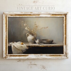 May include: A still life painting in a white frame with gold accents. The painting depicts a white ceramic pitcher with a bouquet of white flowers, a small white teapot, a wooden bowl, and a single orange fruit on a white tablecloth.