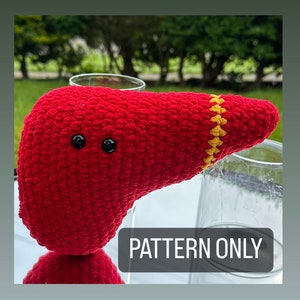 May include: Crochet pattern for a red and yellow liver amigurumi toy. The toy has two black eyes and is sitting on a table next to a glass. The text "PATTERN ONLY" is visible in the image.