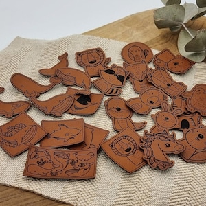 Könnte beinhalten: Eine Sammlung brauner Lederaufnäher mit Tierillustrationen, darunter ein Wal, ein Löwe, ein Fuchs und ein Pinguin. Die Aufnäher haben schwarze Umrisse und Details und sind in verschiedenen Formen und Größen gehalten. Sie liegen auf einem beigen Stoff.