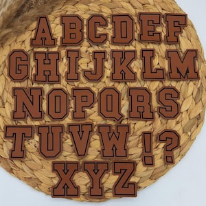 Könnte beinhalten: Braune Lederbuchstaben mit schwarzen Umrandungen. Die Buchstaben sind in alphabetischer Reihenfolge auf einem geflochtenen Stroh-Hintergrund angeordnet.