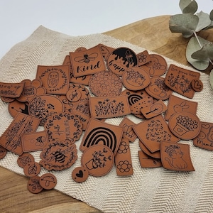 Könnte beinhalten: Eine Sammlung brauner Lederanhänger mit verschiedenen Designs und Texten, darunter "Grow Up", "Flower Power", "Kind", "Happ", "Love you more", "Hello Frühling", "Mumlife" und mehr. Die Anhänger liegen auf einer braunen und weißen gemusterten Oberfläche.