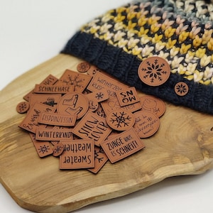 Könnte beinhalten: Eine Sammlung brauner Lederpatches mit verschiedenen winterlichen Wörtern und Illustrationen, darunter "Snow Lover", "Winterkind", "Sweater Weather" und "Zunge raus, es schneit!" Die Patches sind auf einer Holzoberfläche mit einer Strickmütze im Hintergrund angeordnet.