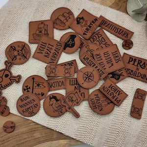 Könnte beinhalten: Eine Sammlung brauner Lederpatches mit verschiedenen Designs und Texten zum Thema Piraten, darunter "Pirate in Ausbildung", "Volle Kraft Vorraus!" und "Bade Wannen-Kapitan".
