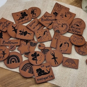 Könnte beinhalten: Eine Sammlung brauner Lederpatches mit schwarzen Illustrationen und Text. Designs umfassen Ballerinas, Einhörner, Regenbogen und Sätze wie "Believe in Magic" und "Ballerina". Die Patches variieren in Form und Größe.