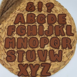 Könnte beinhalten: Braune Buchstaben und Satzzeichen in einer Bubble-Schrift. Die Buchstaben sind auf einem gewebten, kreisförmigen Platzset angeordnet. Die Buchstaben haben eine dunkelbraune Umrandung und sind in einer Serifenschrift.