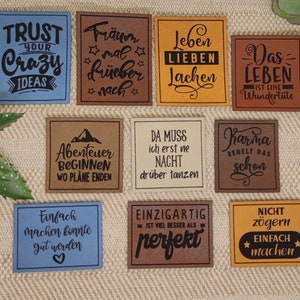 Könnte beinhalten: Neun bunte Leder-Patches mit inspirierenden Zitaten in schwarzer Schrift. Die Zitate lauten: "Vertraue deinen verrückten Ideen", "Träume groß", "Lebe, liebe, lache", "Karma regiert", "Abenteuer beginnen, wo Pläne enden", "Da muss ich erst ne Nacht drüber tanzen", "Einfach machen konnte gut werden", "Einfach machen", und "Das Leben ist eine Wundertüte".