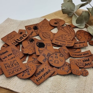 Könnte beinhalten: Eine Sammlung brauner Lederaufnäher mit schwarzen Illustrationen und Text zum Thema Weltraum. Designs umfassen Raketen, Astronauten, Planeten und Sätze wie "UFO" und "Houston, sind wir bald da?"