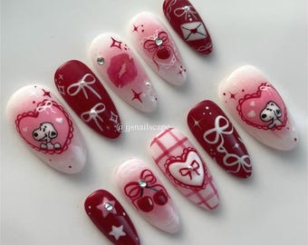 May include: Set of ten red and white almond-shaped press-on nails with various Valentine's Day themed designs, including hearts, bows, cherries, stars, and a cartoon dog. The nails are decorated with glitter and rhinestones. The text "@ijsnailsscape" is written on one of the nails.