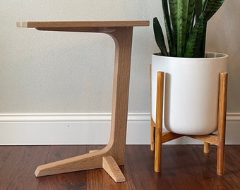 May include: A light wood end table with a curved, open base and a rectangular top. The table is next to a white planter with a green snake plant on a wooden stand.