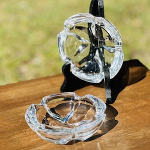 May include: Two clear glass ashtrays with a flower-like design. The ashtrays are on a wooden surface.