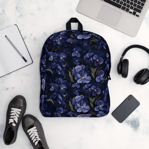 May include: A dark blue backpack with a floral pattern of purple flowers. The backpack is on a white surface with a laptop, headphones, a phone, and two pairs of shoes.