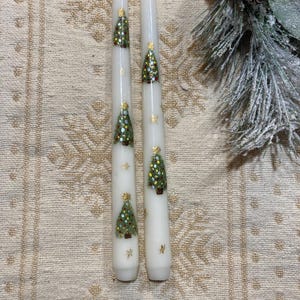 May include: Two white taper candles with green Christmas trees and gold glitter painted on them.