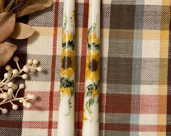 May include: Two white taper candles with hand-painted sunflower designs. The candles are on a plaid fabric background.