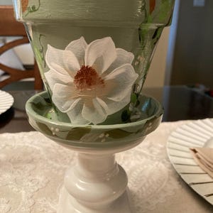 May include: A hand-painted, sage green ceramic flower pot with a matching saucer, set on a white pedestal. The pot features a large white flower with a brown center. Green and brown floral accents adorn the pot's rim.