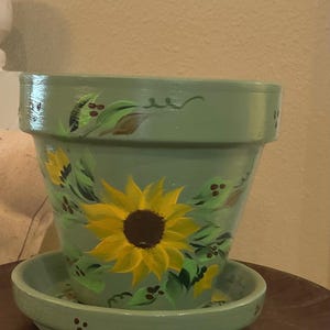 May include: A sage green terracotta plant pot with a matching saucer, featuring hand-painted yellow sunflowers with dark brown centers and green leaves. The design includes small brown dots and vine-like accents painted around the rim and base.