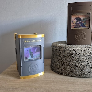 Peut inclure: Deux boîtes de rangement rectangulaires pour cartes avec des fenêtres d'affichage frontales. La boîte de gauche est grise avec des accents dorés et le texte "WorldShaper" au-dessus de la fenêtre. La boîte de droite est marron uni.