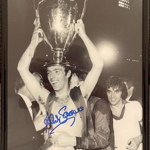May include: A black and white photo of a man holding a trophy above his head. The man is smiling and wearing a jersey. The photo is signed "Alan Sorees".