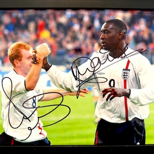 May include: A framed photograph of two soccer players in white jerseys with the England crest. The players are celebrating a goal and have signed their names in black marker. The player on the left is wearing number 8 and the player on the right is wearing number 9.