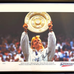 May include: A black and white photo of a woman holding up a gold trophy. The woman is wearing a white tennis outfit and is smiling. The trophy is the Ladies' Winners Plate from the Wimbledon Championships. The photo is signed by Martina Navratilova and includes the text "Sporting Masters" and "03/250".