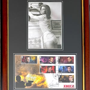 May include: A framed display featuring a black and white photograph of a person in a light-colored top, positioned above a collection of six James Bond postage stamps. The stamps depict various actors and scenes, accompanied by a signature and 007 logo.