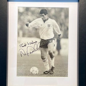 May include: Black and white photograph of a football player in a white shirt with the England flag on the sleeve, dribbling the ball. The player has signed the photo "Best Wishes, N. Phelps".