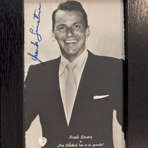 May include: A black and white portrait of Frank Sinatra in a dark suit and light tie, framed in a black wooden frame. The image features a blue ink signature on the left and the text "Frank Sinatra in Das Schicksal hat es so gewollt" below.